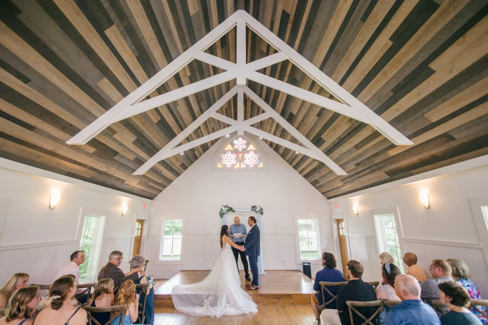 An ultra-wide view of the Grand View Lodge chapel with a vaulted wood-truss ceiling and rose window, where the couple stands at a floral arch exchanging vows as seated guests look on — Tim Larsen Photography, Brainerd Lakes MN