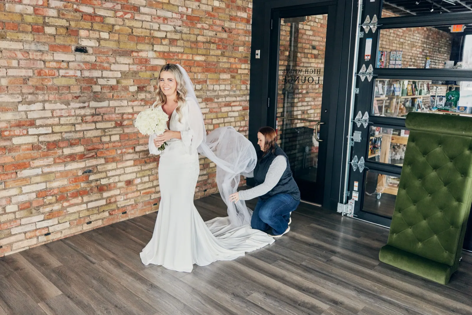 The bride smiles holding a white bouquet while an attendant kneels behind her to arrange the veil train against an exposed brick wall. — Tim Larsen Photography, Brainerd Lakes MN