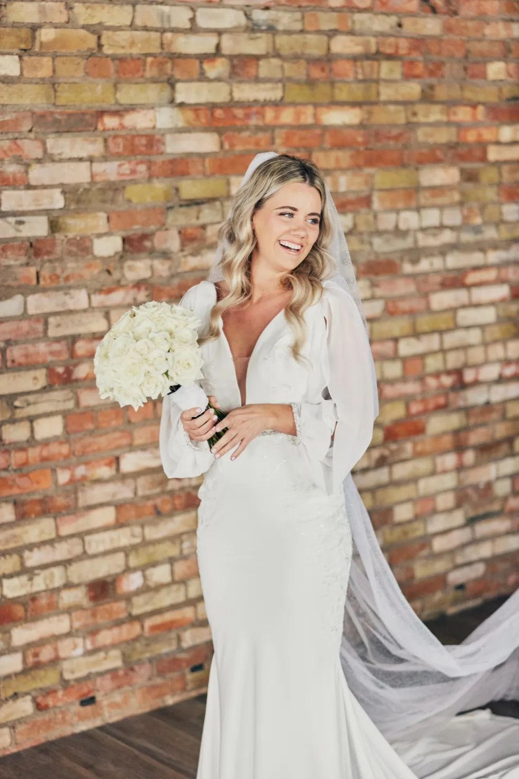 The bride laughs while holding a white rose bouquet, her veil trailing across the floor against an exposed brick wall. — Tim Larsen Photography, Brainerd Lakes MN