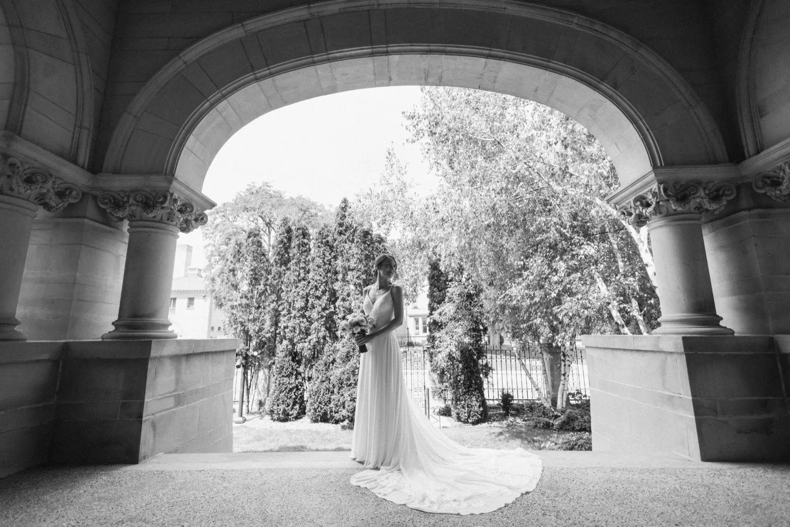 A bride in a flowing gown stands holding her bouquet beneath a massive ornate stone archway with carved columns, her train trailing across the floor. — Tim Larsen Photography, Minneapolis MN