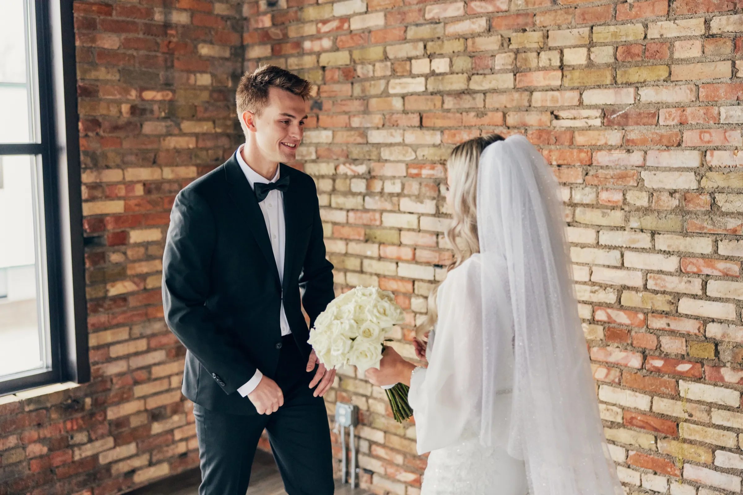 Sam smiles at Madeline during their first look in a room with a tall exposed brick wall and natural window light — Tim Larsen Photography, Brainerd Lakes MN