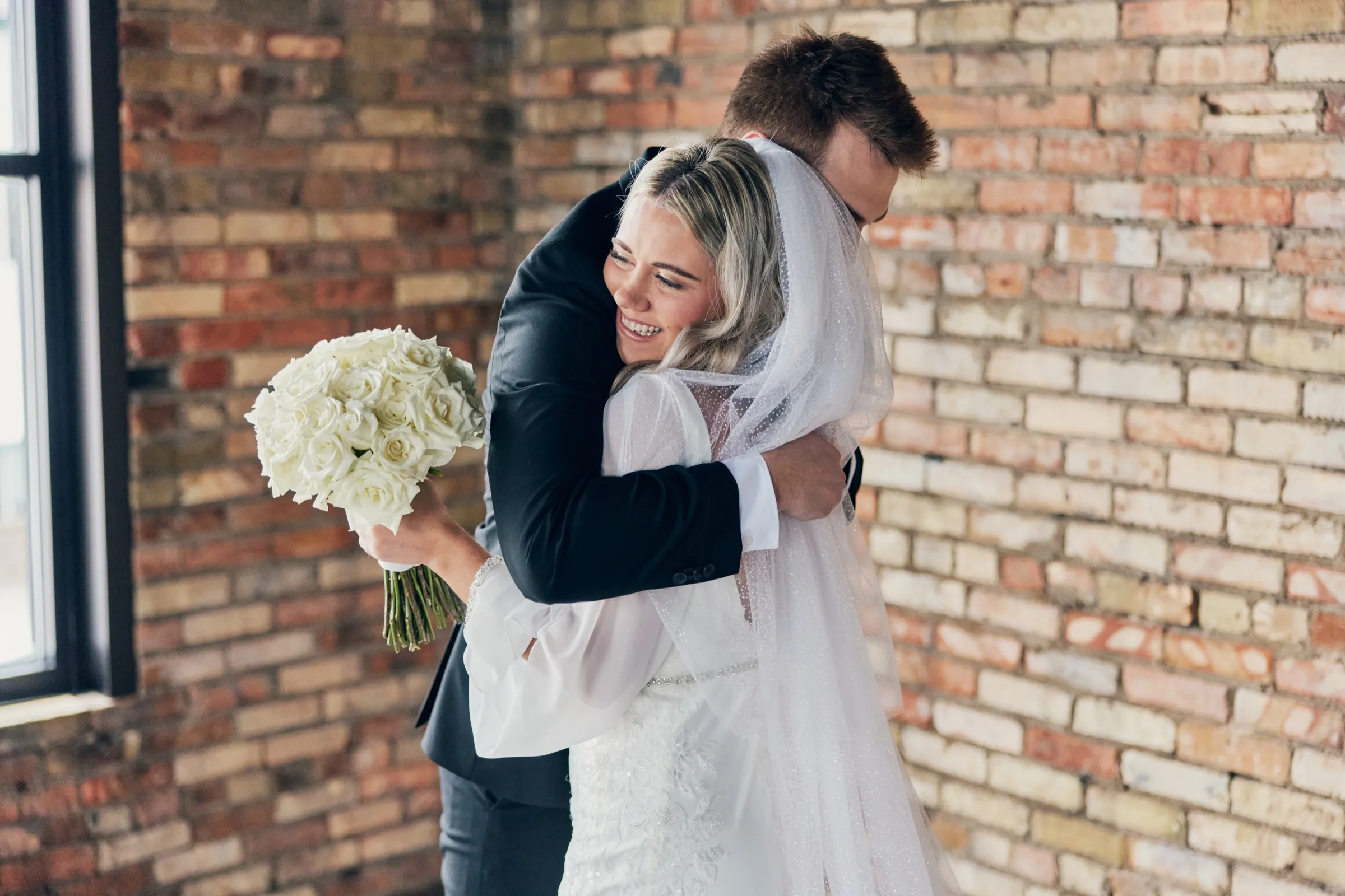 Madeline beams over Sam's shoulder as they embrace at their first look, her white bouquet held out to one side against an exposed brick wall — Tim Larsen Photography, Brainerd Lakes MN