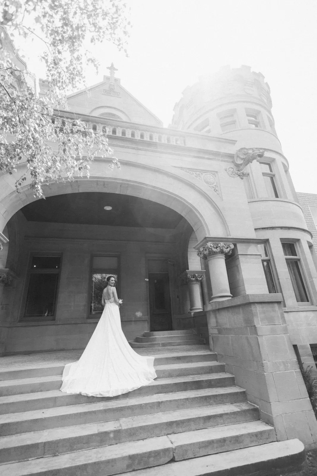 A bride glances over her bare shoulder while her gown's long train spills down the stone steps of a grand arched mansion entrance, photographed in black and white. — Tim Larsen Photography, Minneapolis MN