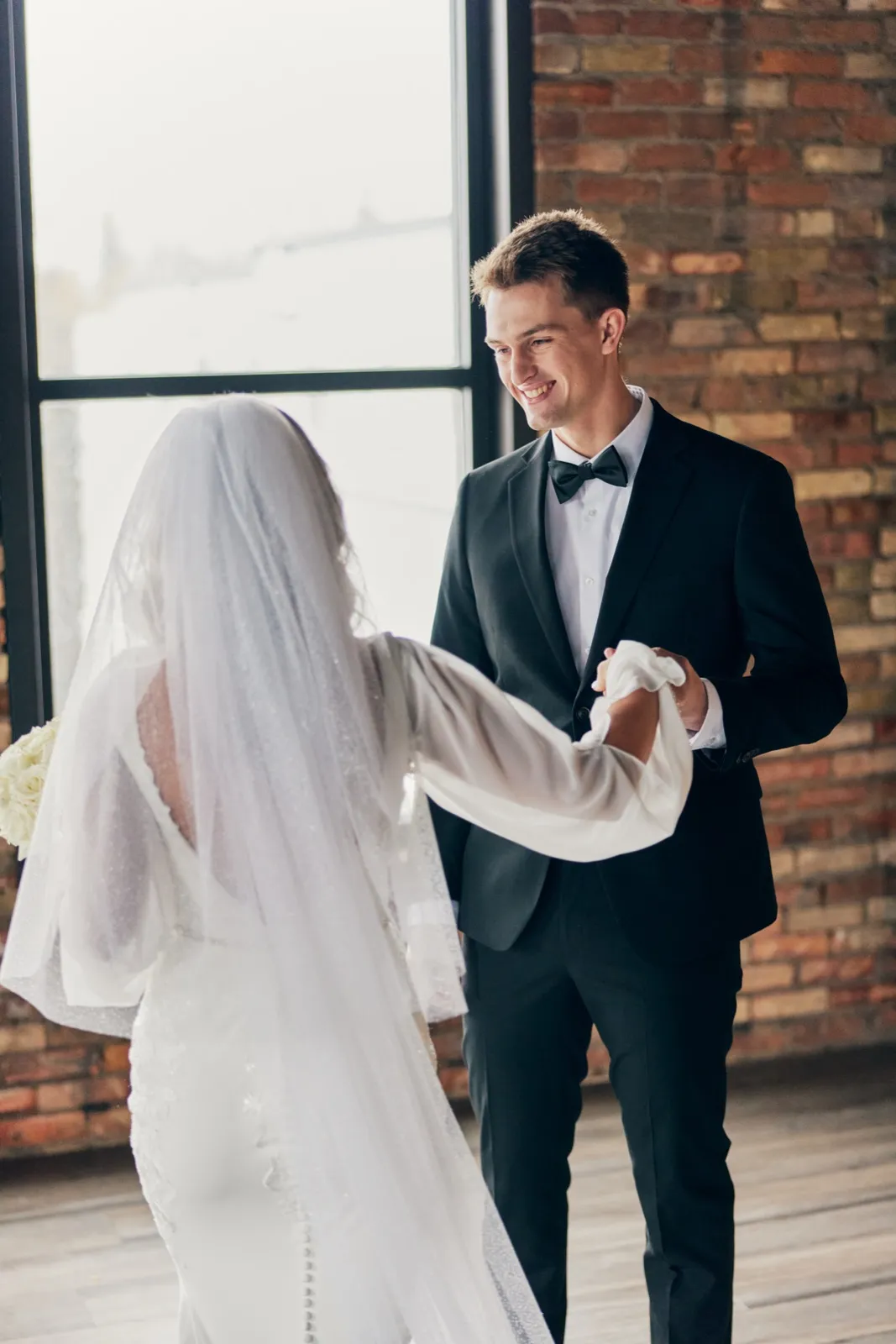 The groom smiles as he reaches for the bride's hands during their first look in a brick-walled room with tall windows behind her glowing veil. — Tim Larsen Photography, Brainerd Lakes MN