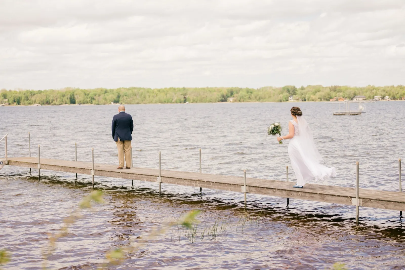 A groom waits with his back turned at the end of a long lake dock as the bride approaches behind him with her bouquet, just before their first look — Tim Larsen Photography, Brainerd Lakes MN