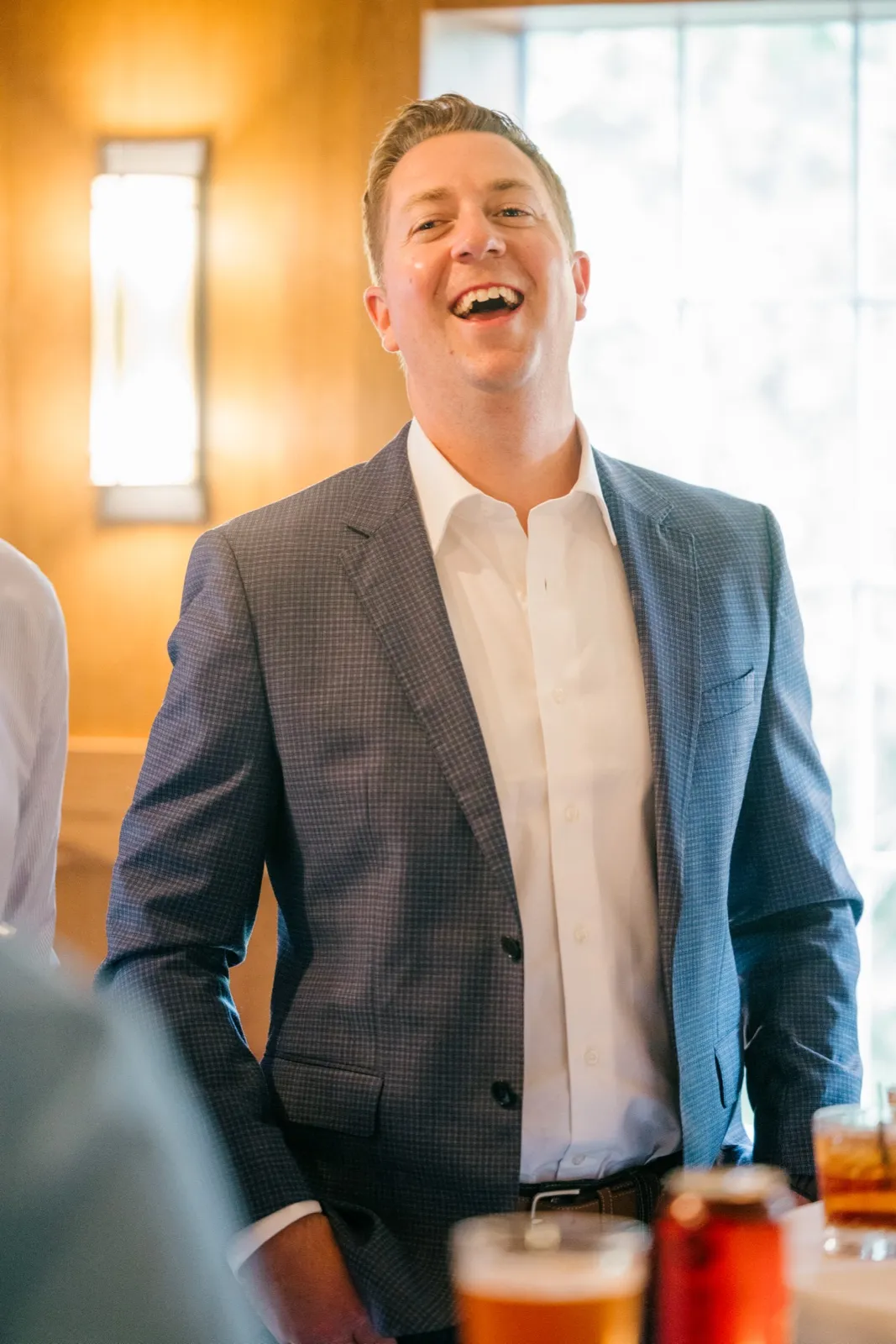 The groom laughs openly in a checked blue suit jacket while standing in a warmly lit room with a tall window behind him — Tim Larsen Photography, Brainerd Lakes MN