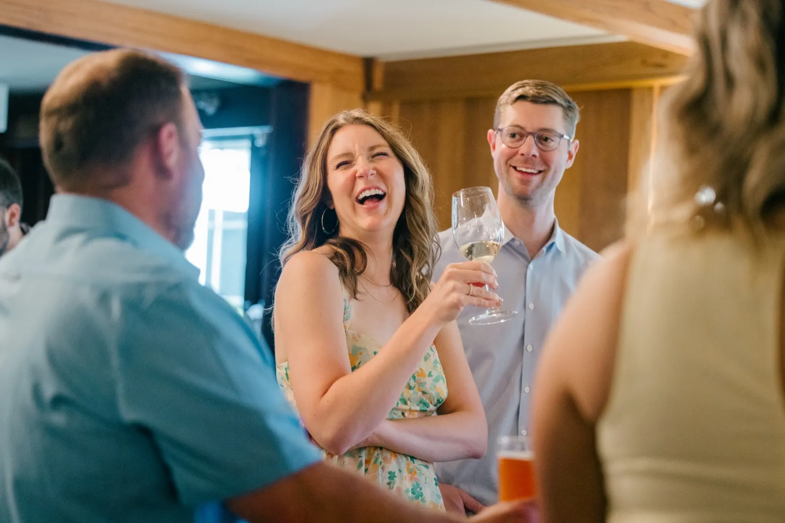 A guest in a floral dress laughs openly while holding a glass of white wine, framed between two other guests in a wood-paneled lodge room — Tim Larsen Photography, Brainerd Lakes MN