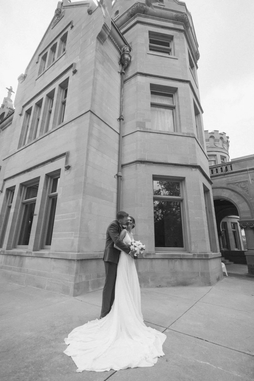 The bride and groom embrace at the base of an ornate stone tower at the American Swedish Institute, her gown's train fanning across the pavement in black and white. — Tim Larsen Photography, Minneapolis MN