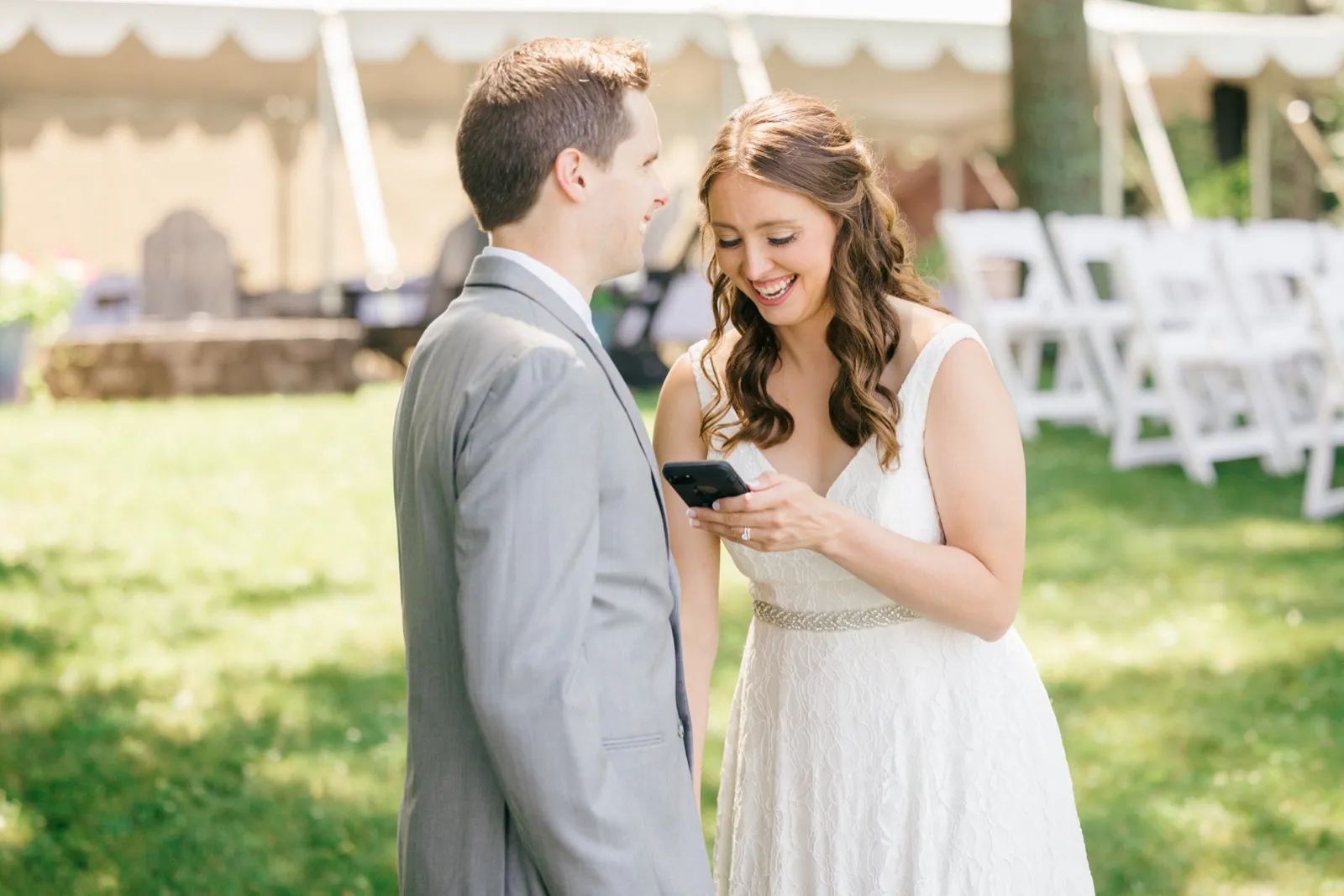 The bride laughs while looking at a phone she holds, as the groom watches her with a smile on a sunlit lawn near the reception tent. — Tim Larsen Photography, Brainerd Lakes MN