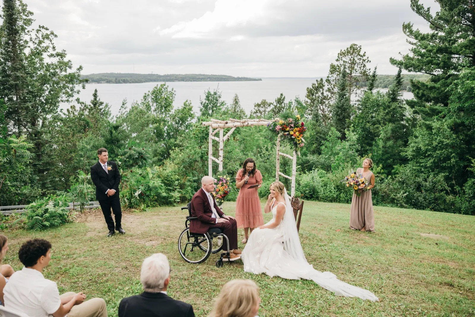 The bride kneels in her gown beside the groom in his wheelchair under a birch arch with wildflowers as the officiant reads, with a lake and tree line behind them. — Tim Larsen Photography, Brainerd Lakes MN