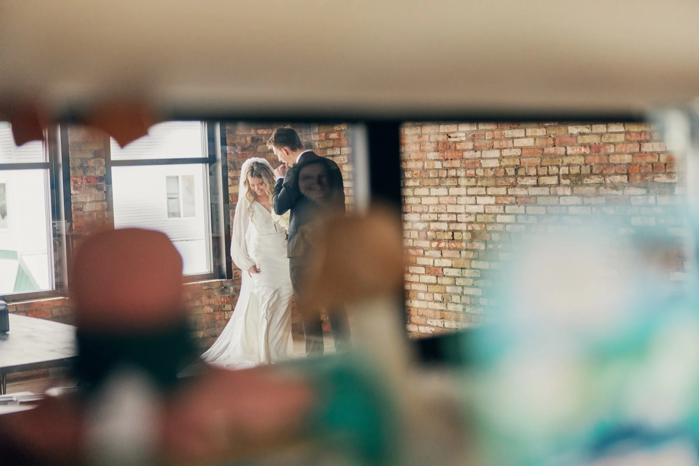 Sam kisses Madeline's hand as they stand together against an exposed brick wall, framed through blurred foreground objects — Tim Larsen Photography, Brainerd Lakes MN