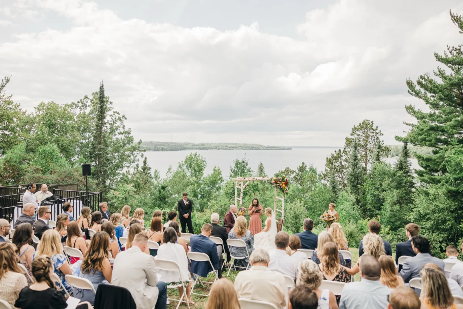 A wide view of an outdoor ceremony on a lawn above a large lake, with the couple at a floral arch facing the officiant and guests seated in rows under a cloudy sky. — Tim Larsen Photography, Brainerd Lakes MN