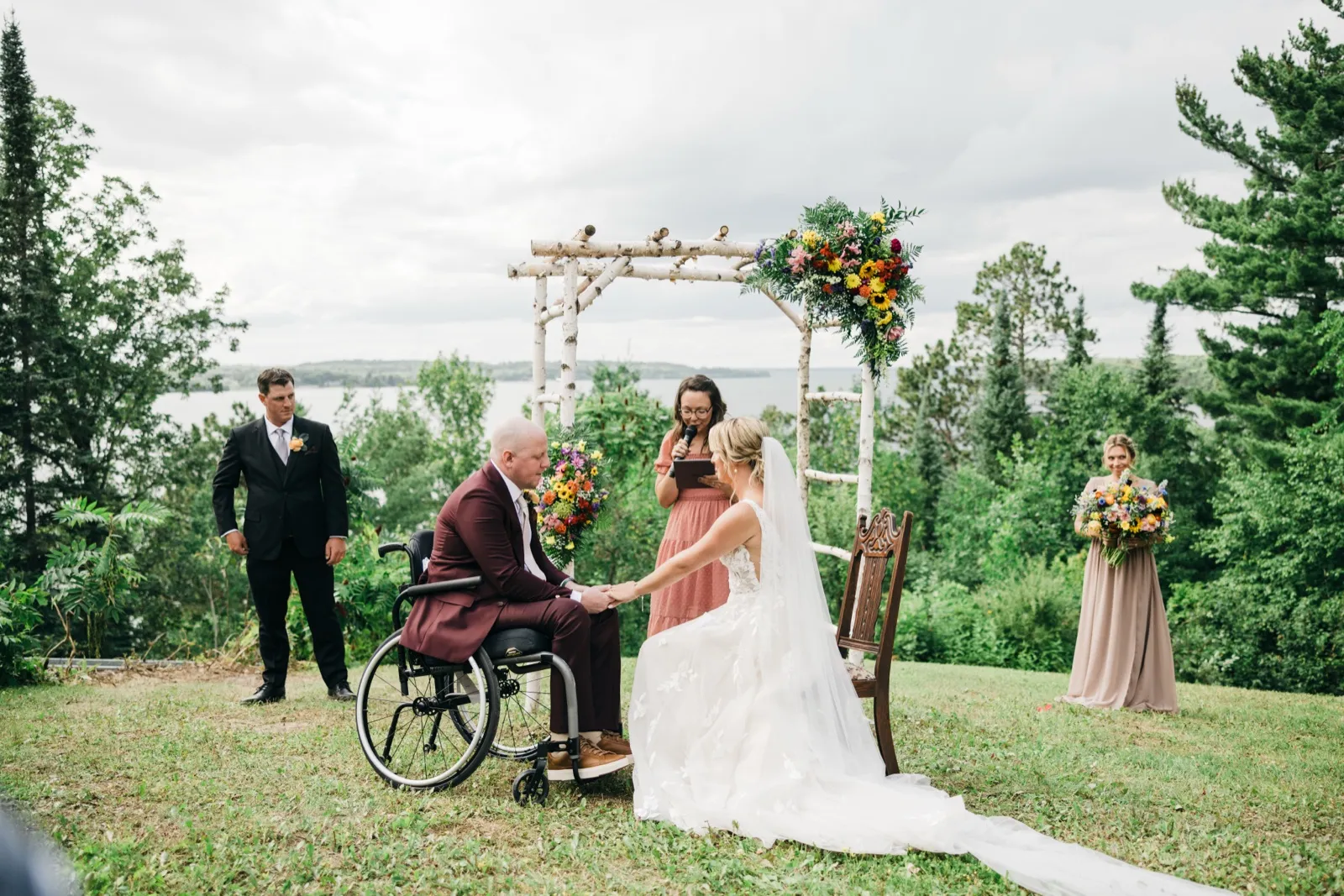 The bride stands holding the groom's hands as he sits in his wheelchair beneath a birch ceremony arch, with the officiant reading vows and the lake visible behind them. — Tim Larsen Photography, Brainerd Lakes MN
