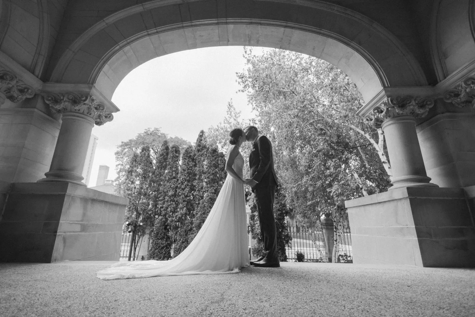 The bride and groom share a kiss beneath a grand stone archway flanked by ornate columns, with the bride's long train trailing across the stone floor. — Tim Larsen Photography, Minneapolis MN