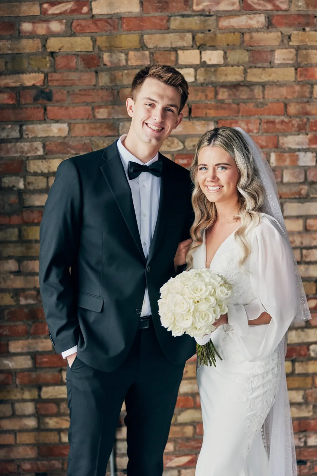 The bride and groom stand close together smiling at the camera against an exposed red brick wall, the bride holding a large white rose bouquet. — Tim Larsen Photography, Brainerd Lakes MN