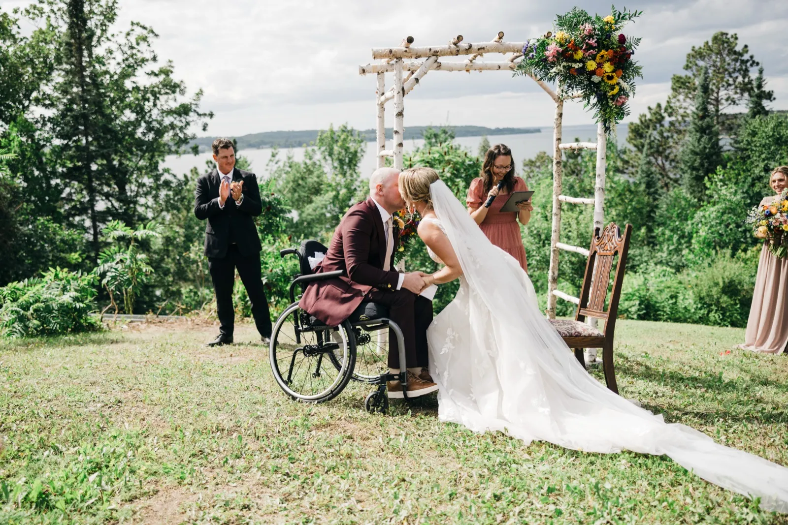 The groom in a wheelchair and the bride share a kiss beneath a birch ceremony arch with a lake horizon behind them, as the officiant and a guest look on. — Tim Larsen Photography, Brainerd Lakes MN