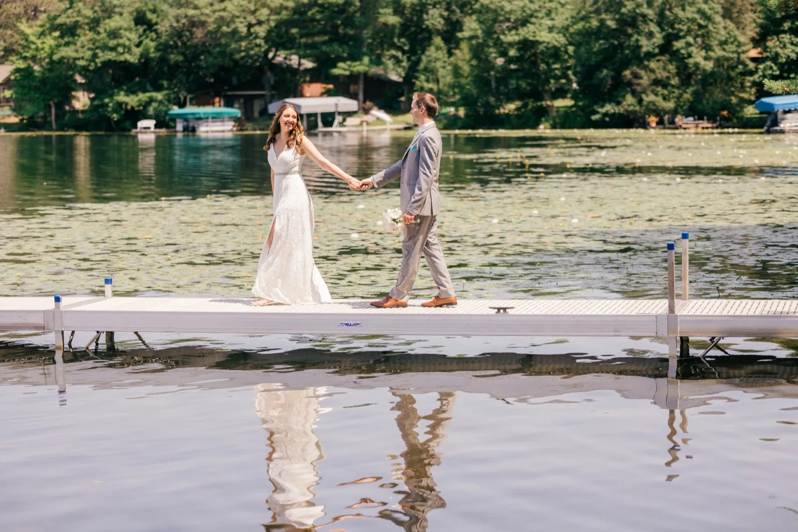The bride looks back at the camera while holding the groom's hand as they walk along a dock, their figures mirrored in the still lake water below. — Tim Larsen Photography, Brainerd Lakes MN