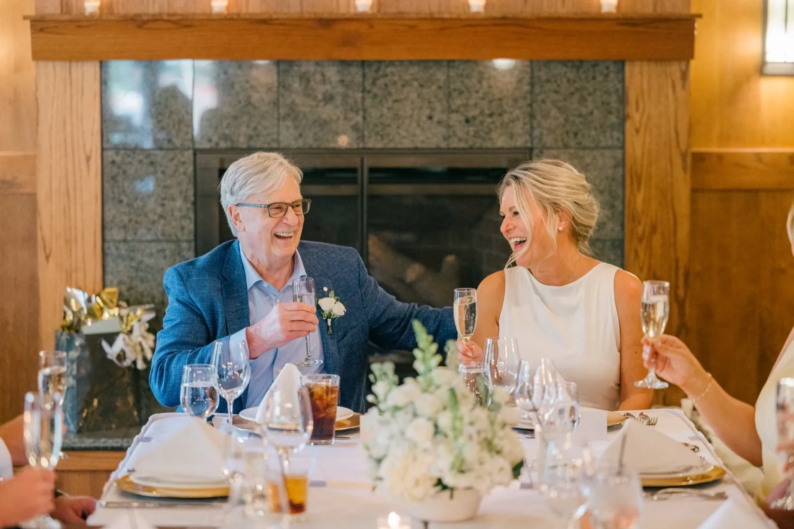 The bride and an older man in a blue suit laugh together while raising champagne flutes at a candlelit dinner table in front of a stone fireplace at Grand View Lodge — Tim Larsen Photography, Brainerd Lakes MN