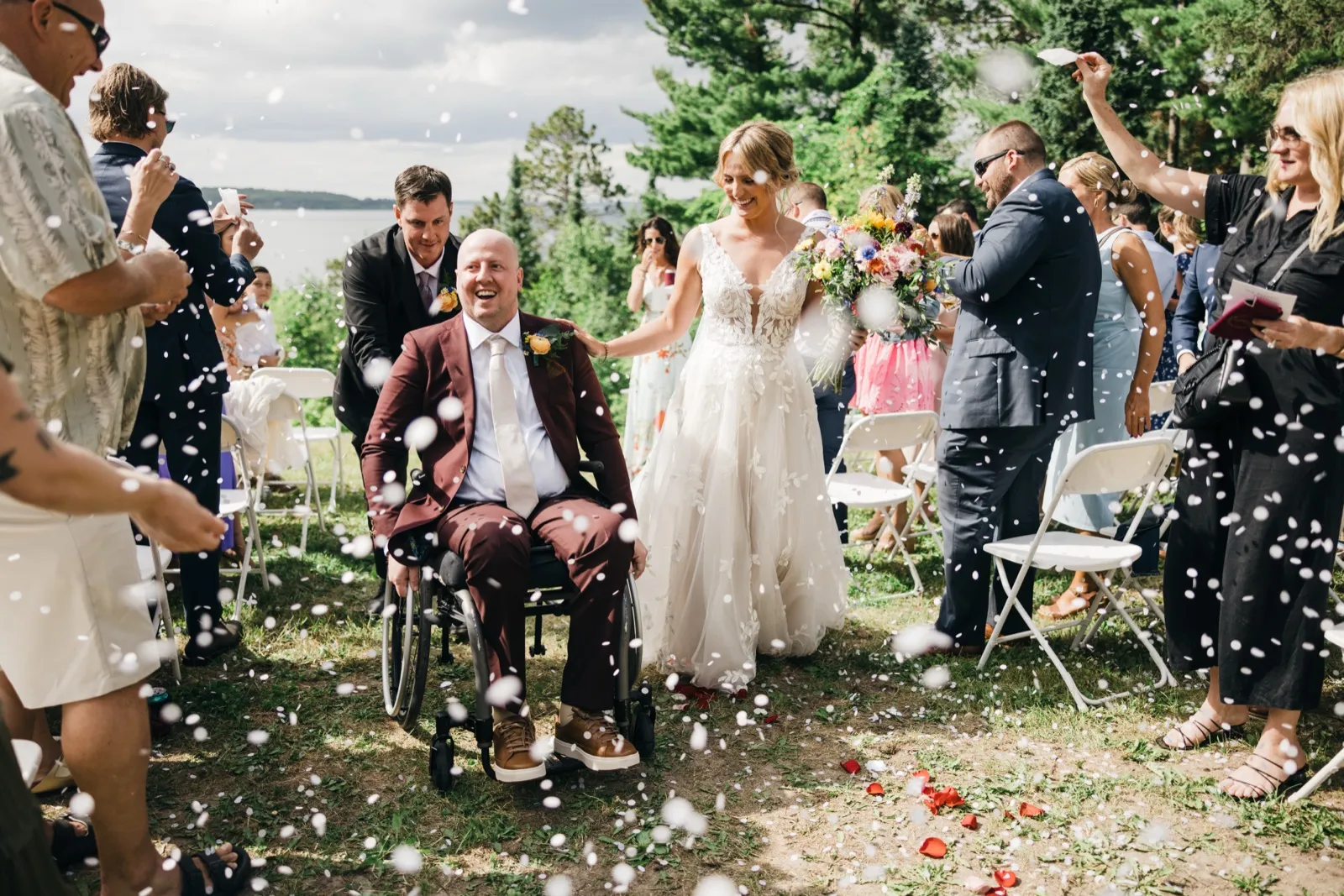 The groom in a wheelchair and the bride holding a colorful bouquet move down the aisle as guests shower them with flower petals at a lakeside ceremony. — Tim Larsen Photography, Brainerd Lakes MN