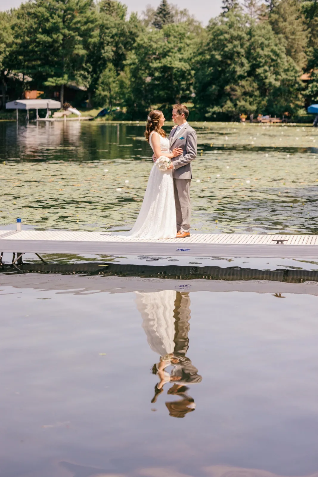 The bride and groom stand together on a wooden dock holding each other, their full figures mirrored in the still lake water below. — Tim Larsen Photography, Brainerd Lakes MN