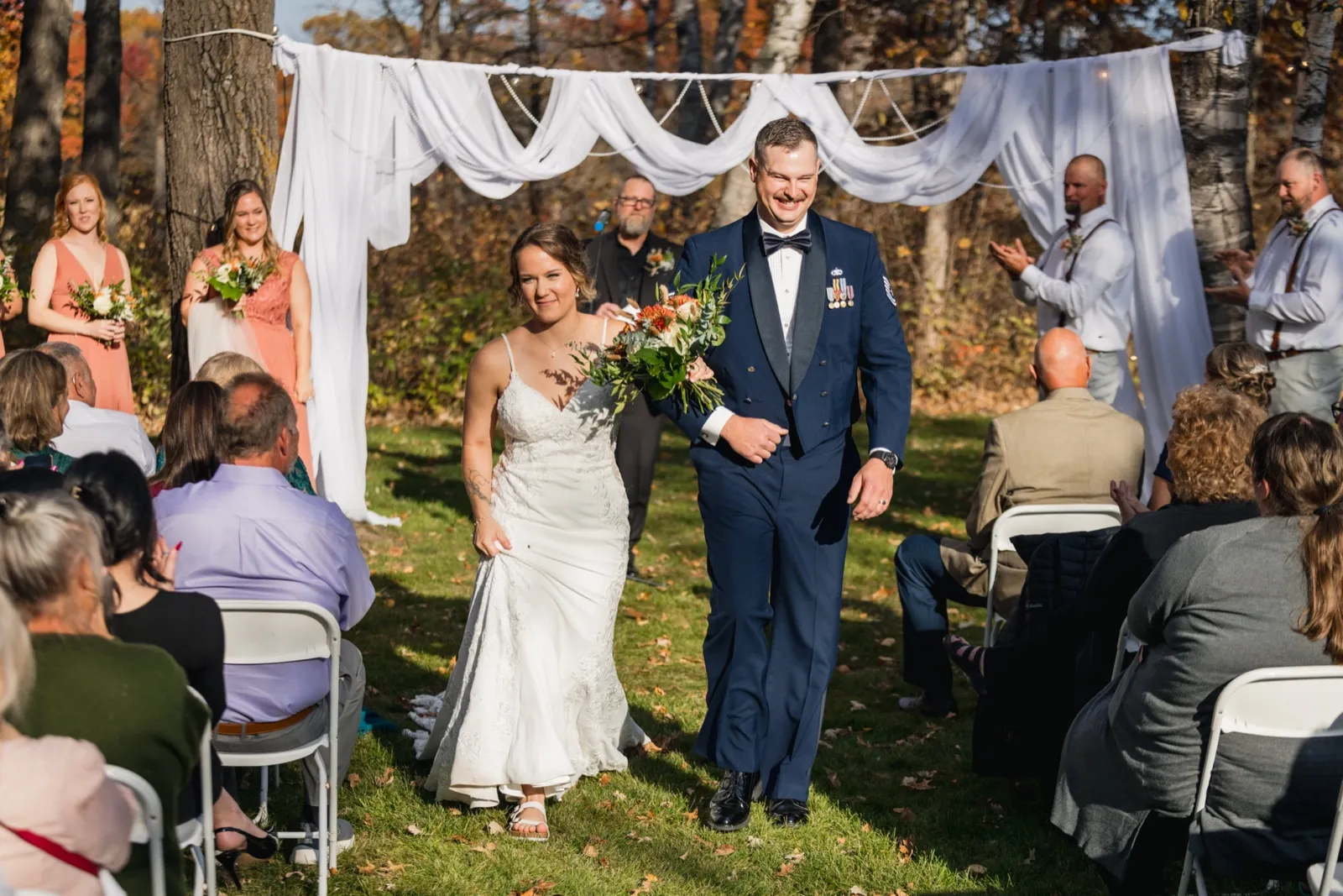 The bride and groom walk back down the aisle together after the ceremony, framed by a white draped arch and autumn trees, with guests and bridal party watching on either side — Tim Larsen Photography, Brainerd Lakes MN