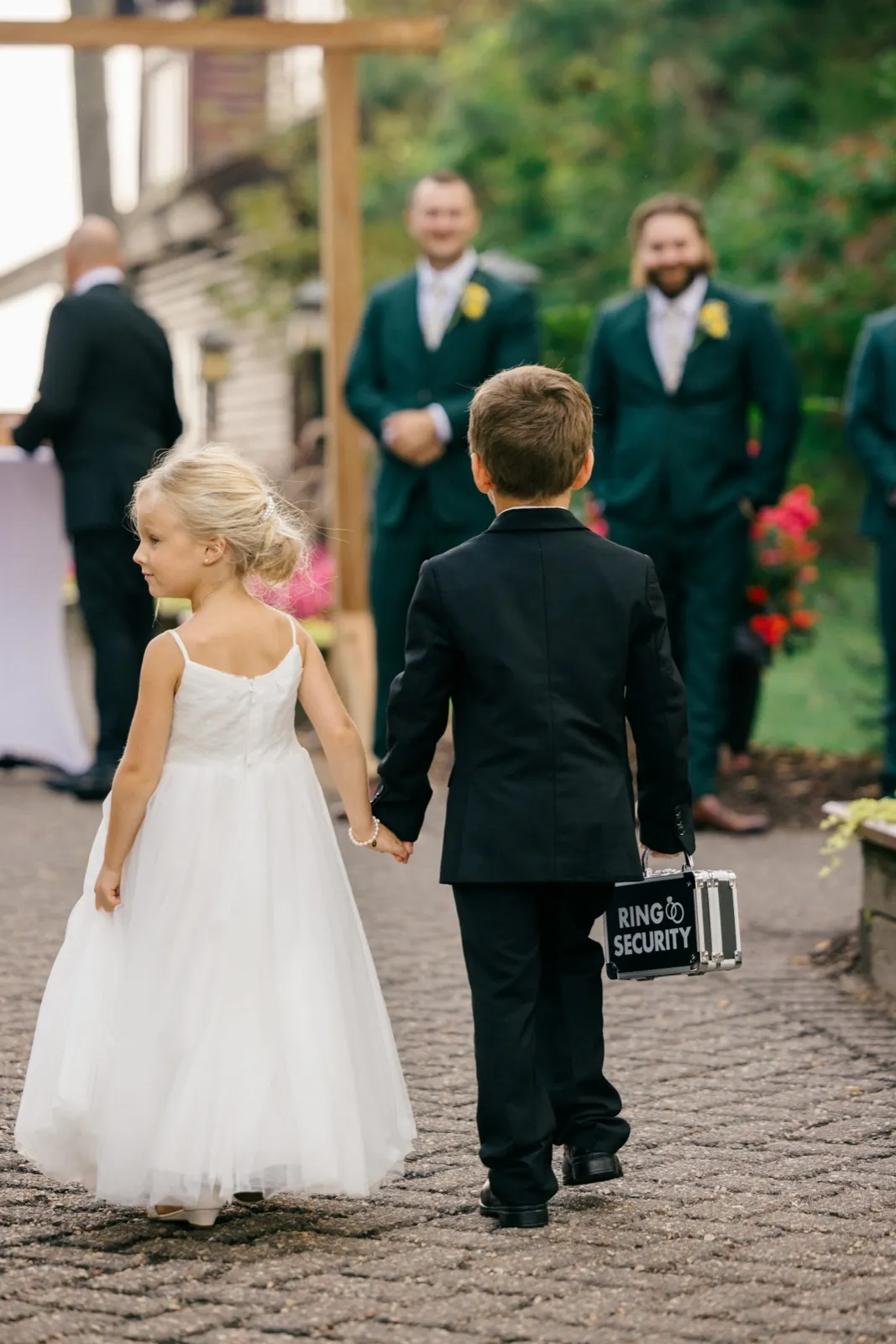 A flower girl in a white tulle dress holds hands with a ring bearer carrying a 'Ring Security' box as they walk down the aisle toward the waiting groom and groomsmen