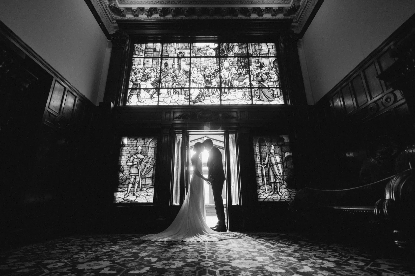 The bride and groom embrace in silhouette beneath a towering stained glass window in the grand entry hall of the American Swedish Institute. — Tim Larsen Photography, Minneapolis MN