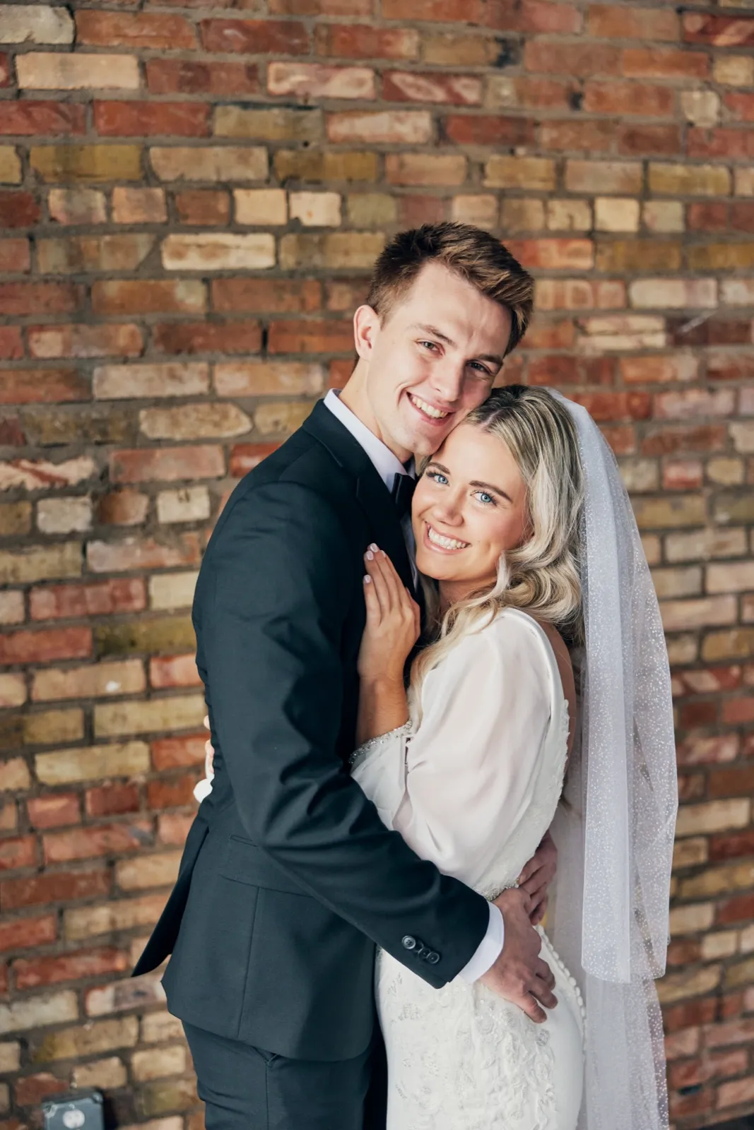 The bride leans her head against the groom's shoulder as both smile toward the camera in front of an exposed brick wall, her long beaded veil cascading down her right side. — Tim Larsen Photography, Brainerd Lakes MN