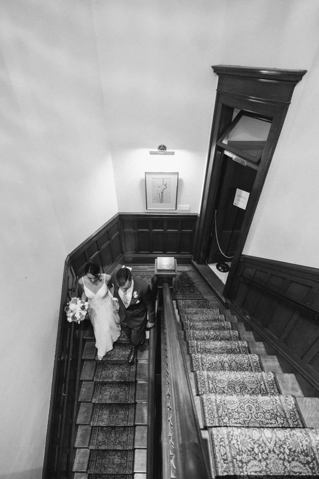 A high-angle black-and-white view of the bride and groom descending a wood-paneled staircase with a patterned runner inside the American Swedish Institute. — Tim Larsen Photography, Minneapolis MN