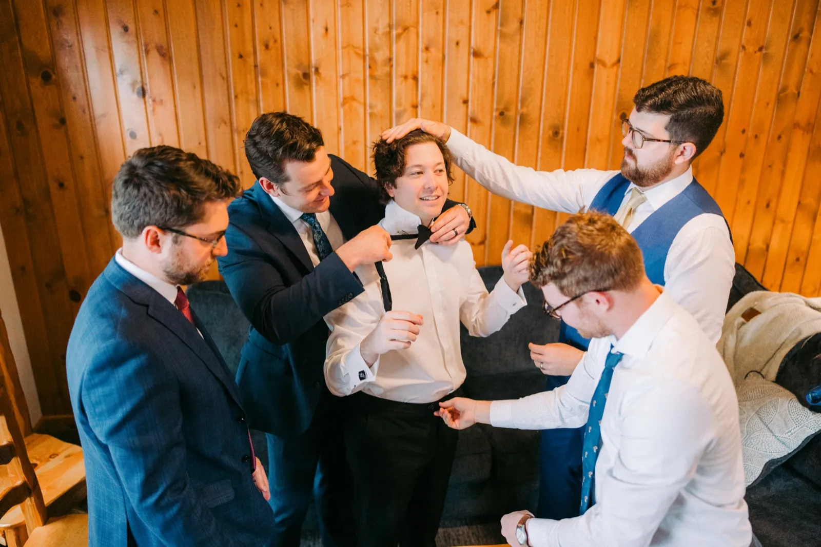 From a high angle, four groomsmen surround the groom in a wood-paneled cabin room, helping him tie his bow tie and smooth his hair — Tim Larsen Photography, Brainerd Lakes MN