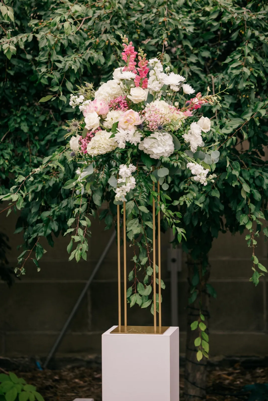 A tall floral arrangement of pink and white blooms with trailing greenery sits atop a gold stand and white pedestal, set against a backdrop of dense green foliage. — Tim Larsen Photography, Minneapolis MN
