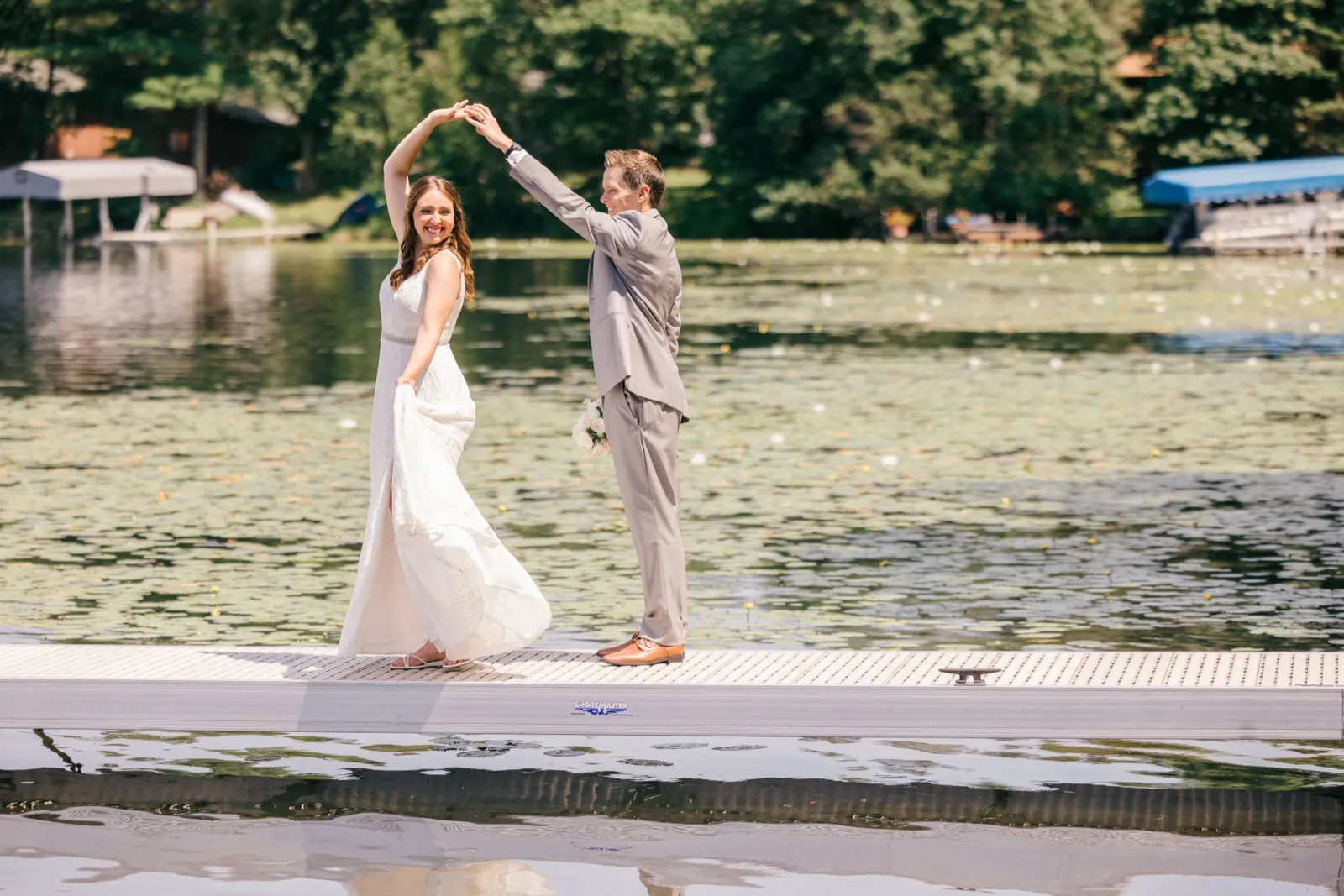 The groom twirls the bride on a lakeside dock as her dress sweeps wide and both of them laugh, with green trees and lily pads filling the water behind them. — Tim Larsen Photography, Brainerd Lakes MN