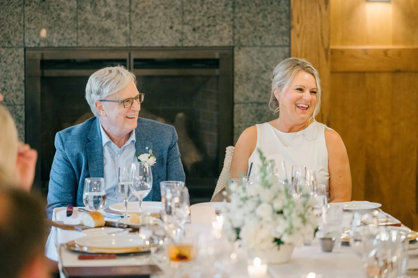 The bride laughs openly beside an older male guest in a blue jacket as they sit at a reception table set with white florals in front of a stone fireplace — Tim Larsen Photography, Brainerd Lakes MN