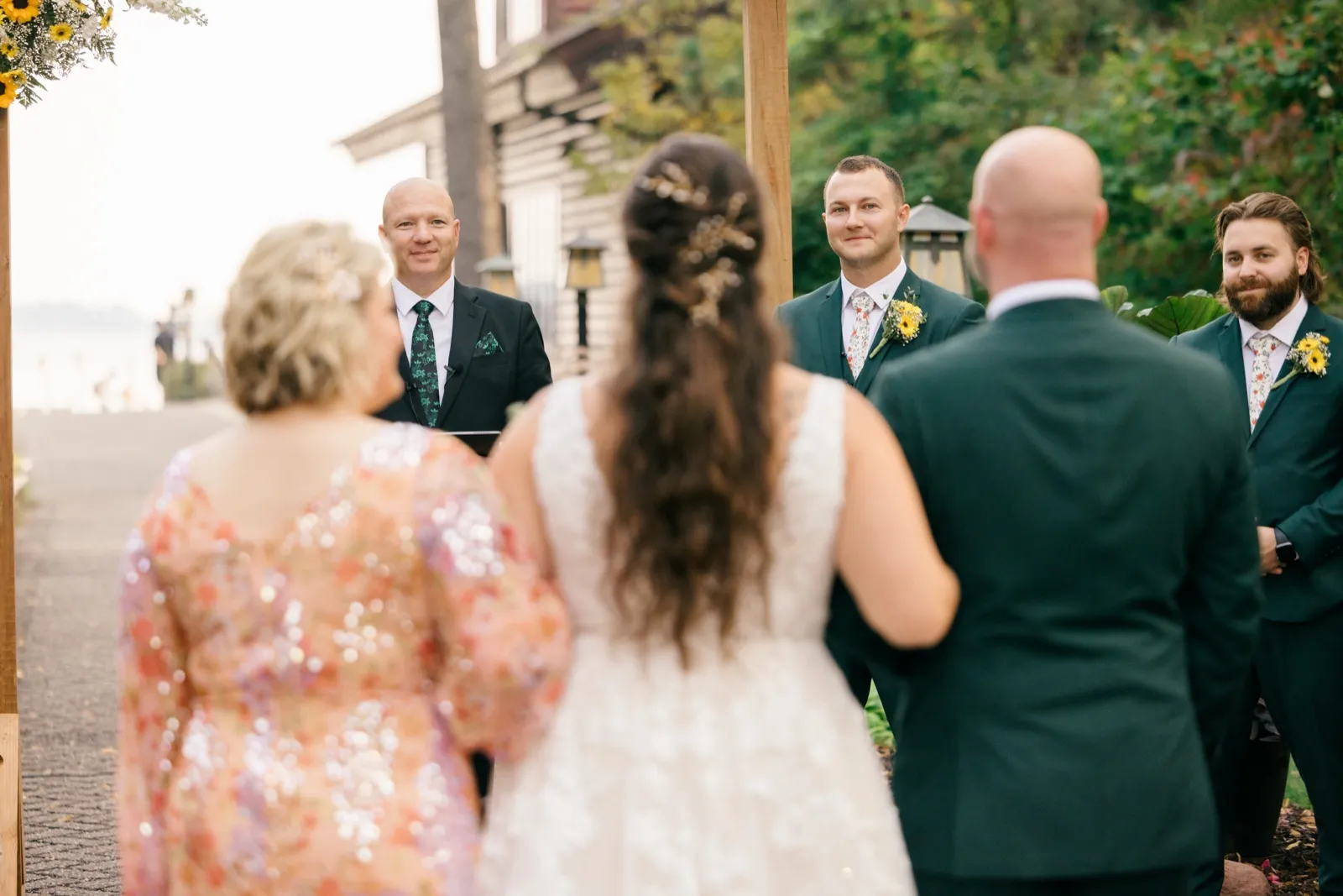 Eric smiles toward Chelsea from beneath a wooden ceremony arch as she stands with her mother in the foreground, surrounded by groomsmen — Tim Larsen Photography, Brainerd Lakes MN