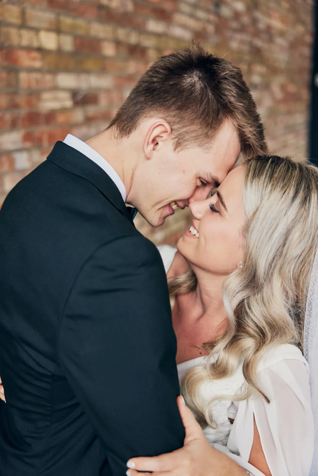 The bride laughs with her eyes closed as she presses her forehead against the groom's nose in front of an exposed brick wall. — Tim Larsen Photography, Brainerd Lakes MN