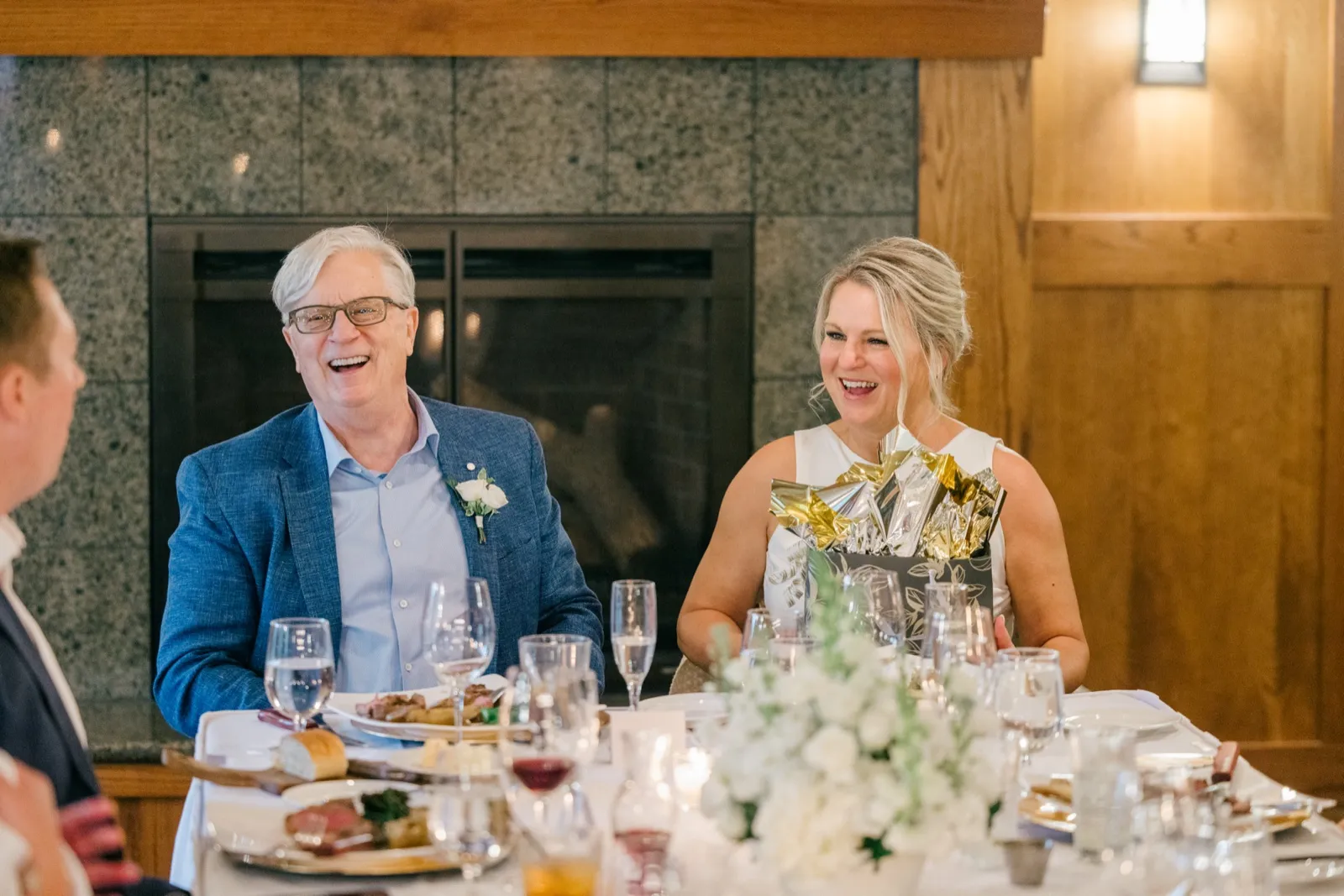 Two seated wedding guests burst into simultaneous laughter at a reception table, with a tall stone fireplace and white florals framing the scene — Tim Larsen Photography, Brainerd Lakes MN