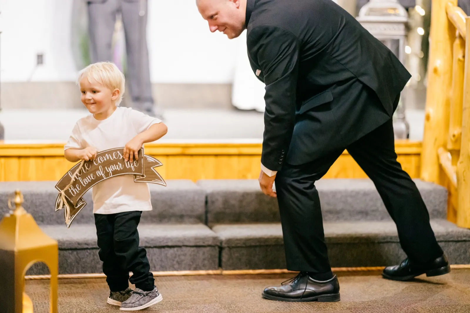 A small ring bearer grins as he carries a hand-lettered sign reading 'here comes the love of your life' down the aisle while Jesse bends forward in greeting — Tim Larsen Photography, Brainerd Lakes MN