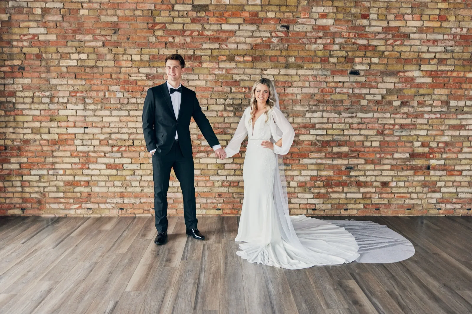 The bride and groom hold hands and smile at the camera in front of a large exposed brick wall, with the bride's long train fanned out across the wood floor. — Tim Larsen Photography, Brainerd Lakes MN
