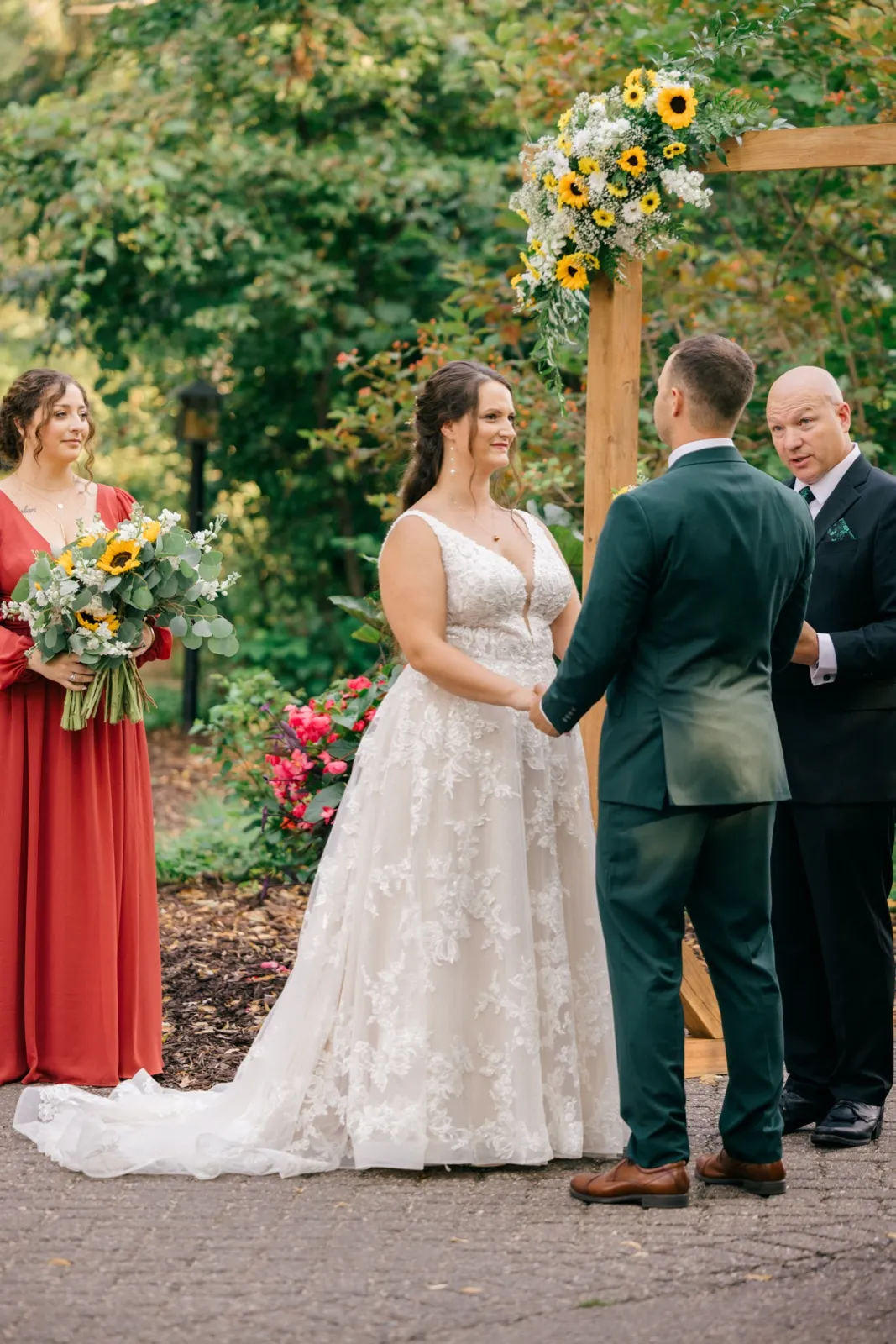 The bride holds her groom's hands beneath a wooden arch decorated with sunflowers as a bridesmaid and officiant look on at the outdoor ceremony