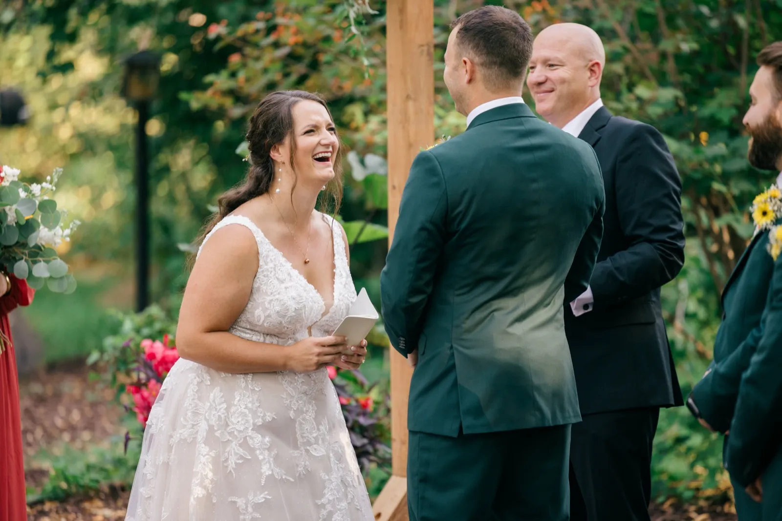 Chelsea bursts into laughter while holding her vow book during the outdoor garden ceremony, with Eric and a smiling officiant in the foreground — Tim Larsen Photography, Brainerd Lakes MN