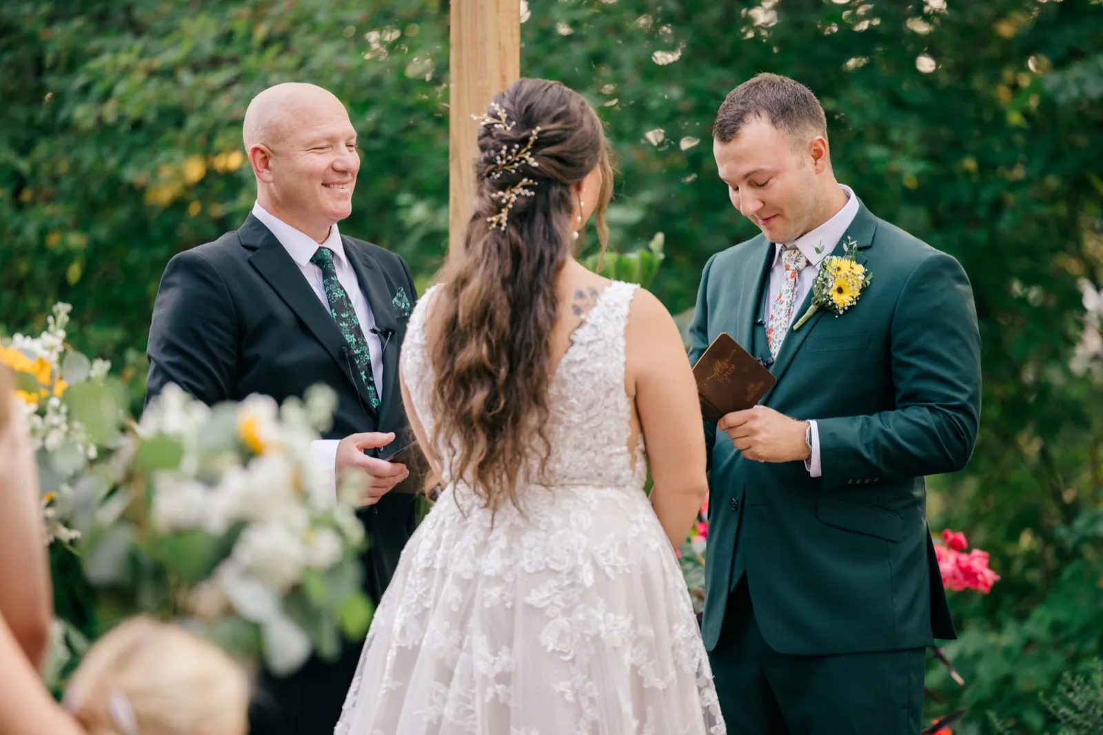 The officiant smiles warmly at Chelsea while Eric reads from a leather vow book during their outdoor ceremony framed by greenery — Tim Larsen Photography, Brainerd Lakes MN