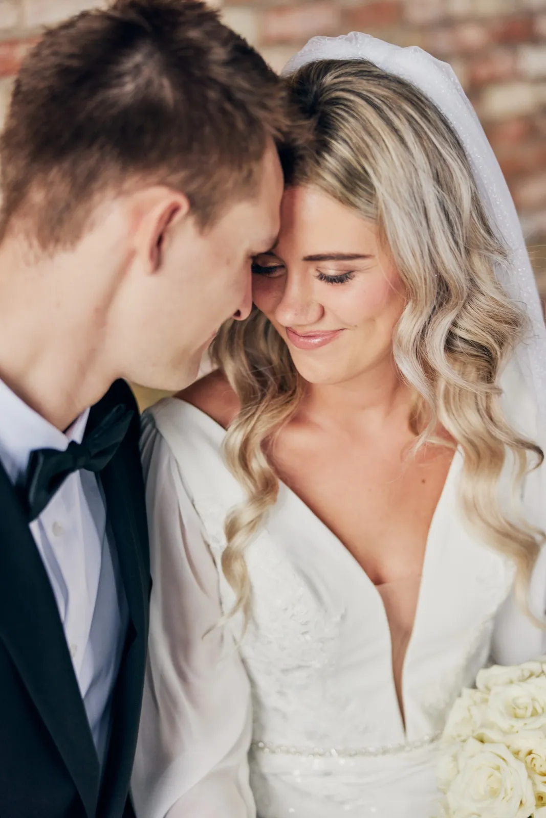 The bride smiles softly with her eyes closed as the groom leans his forehead against hers in front of a brick wall. — Tim Larsen Photography, Brainerd Lakes MN