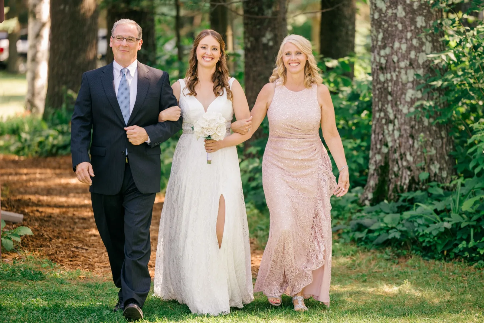 The bride walks down a wooded path toward her ceremony, flanked by her father in a navy suit and her mother in a blush gown, all three smiling. — Tim Larsen Photography, Brainerd Lakes MN