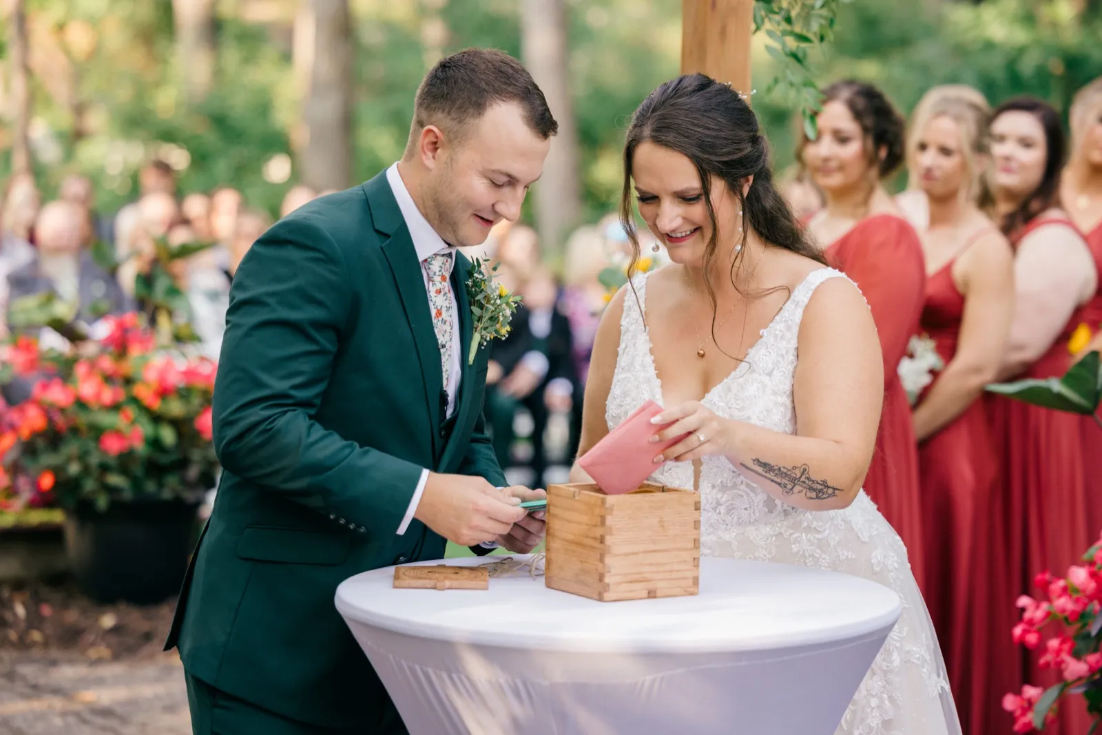 Chelsea and Eric smile down at letters they are placing into a wooden time-capsule box during their outdoor forest ceremony, with bridesmaids in red looking on — Tim Larsen Photography, Brainerd Lakes MN