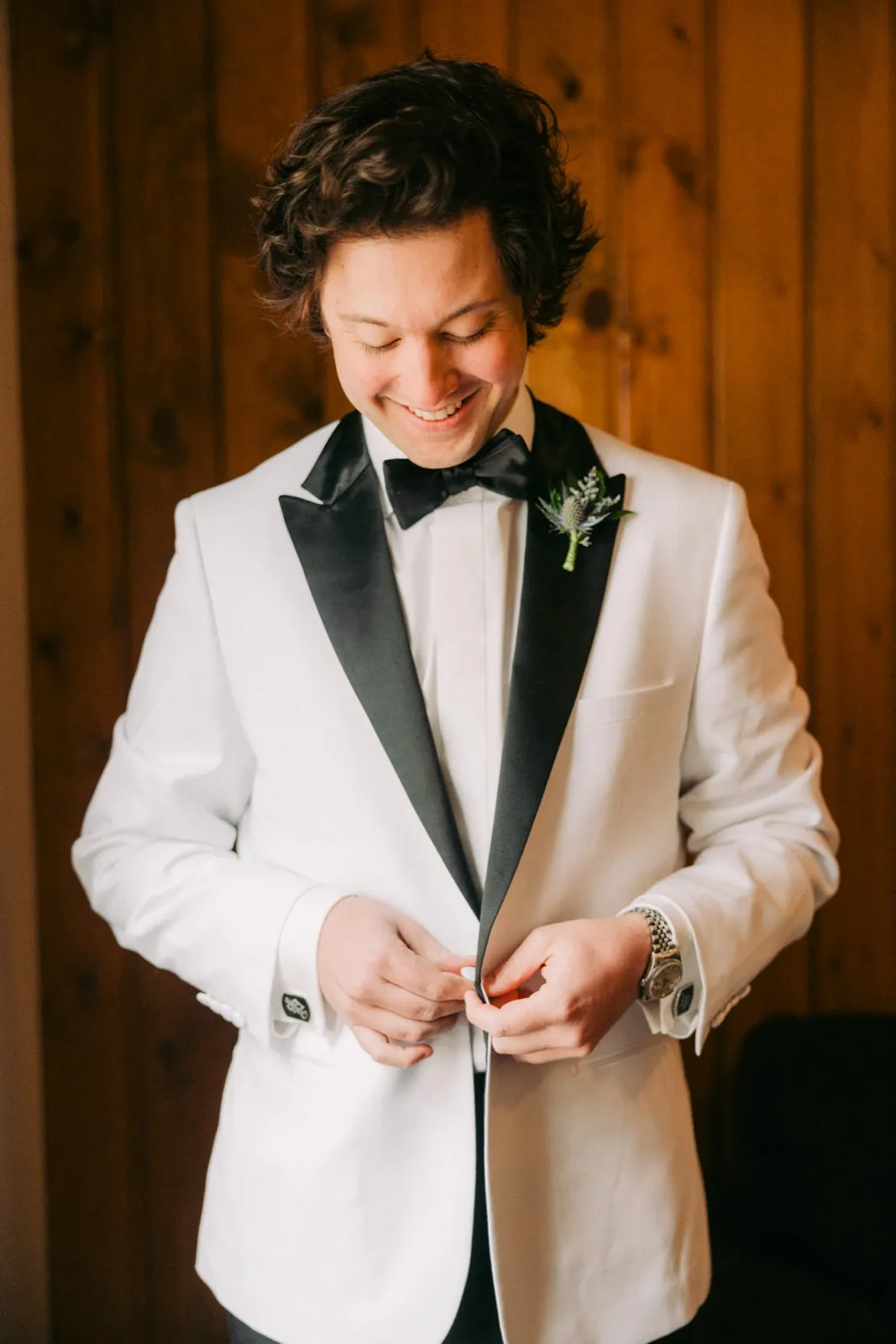 The groom smiles down as he buttons his white tuxedo jacket in a wood-paneled getting-ready room — Tim Larsen Photography, Brainerd Lakes MN