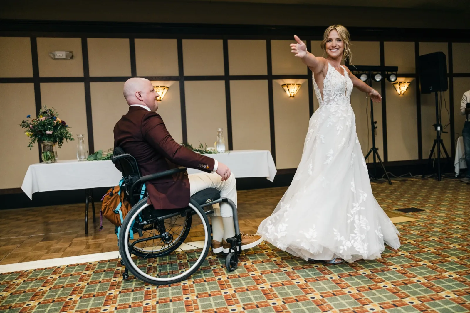 The bride twirls with arms outstretched and a wide smile as the groom watches from his wheelchair on a patterned ballroom dance floor. — Tim Larsen Photography, Brainerd Lakes MN