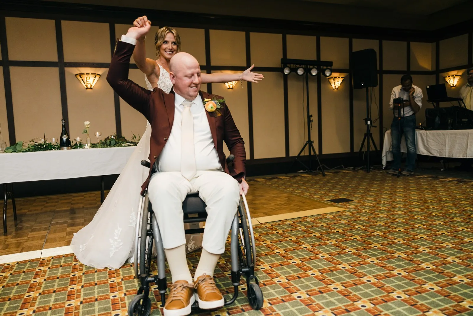 Joe throws an arm out wide from his wheelchair, pointing across the dance floor at Jenna, who raises a fist behind him with a grin during their first dance. — Tim Larsen Photography, Brainerd Lakes MN
