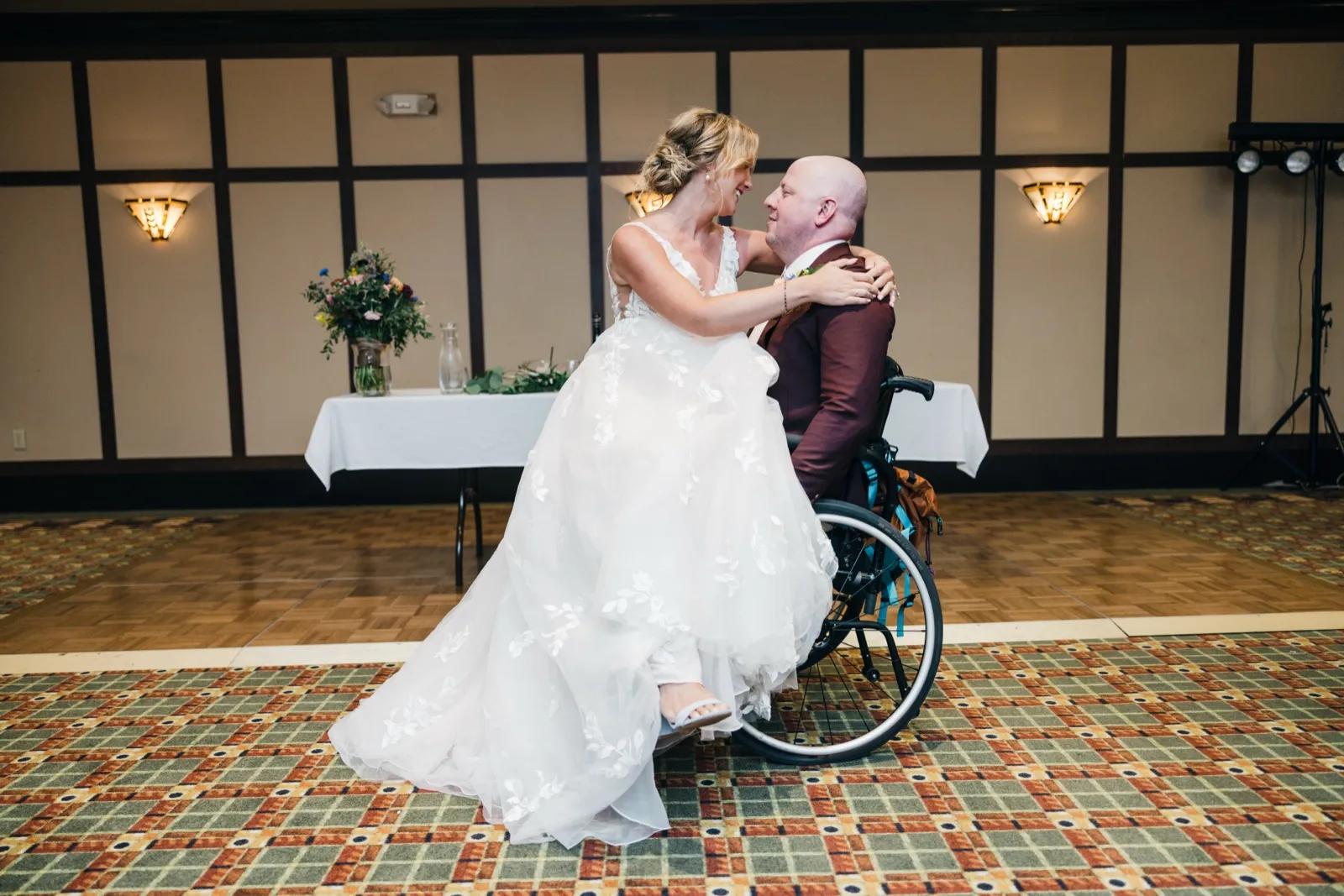 The bride leans into the groom, who is seated in a wheelchair, as they share their first dance on a patterned ballroom floor with the sweetheart table behind them. — Tim Larsen Photography, Brainerd Lakes MN