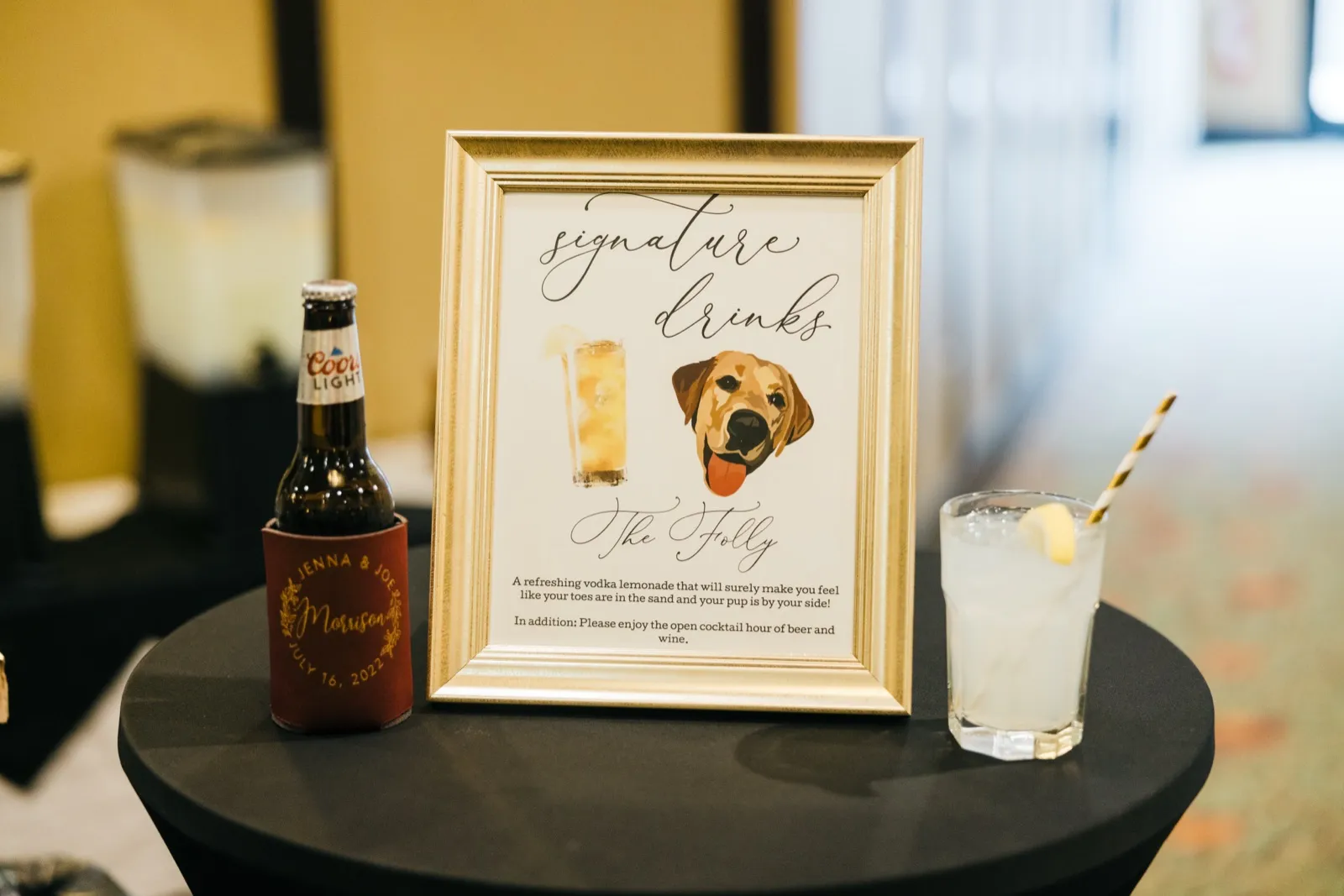 A framed signature drinks sign featuring an illustrated dog named The Folly sits on a cocktail table beside a Coors Light in a personalized Jenna & Joe koozie and a lemonade cocktail with a striped straw. — Tim Larsen Photography, Brainerd Lakes MN