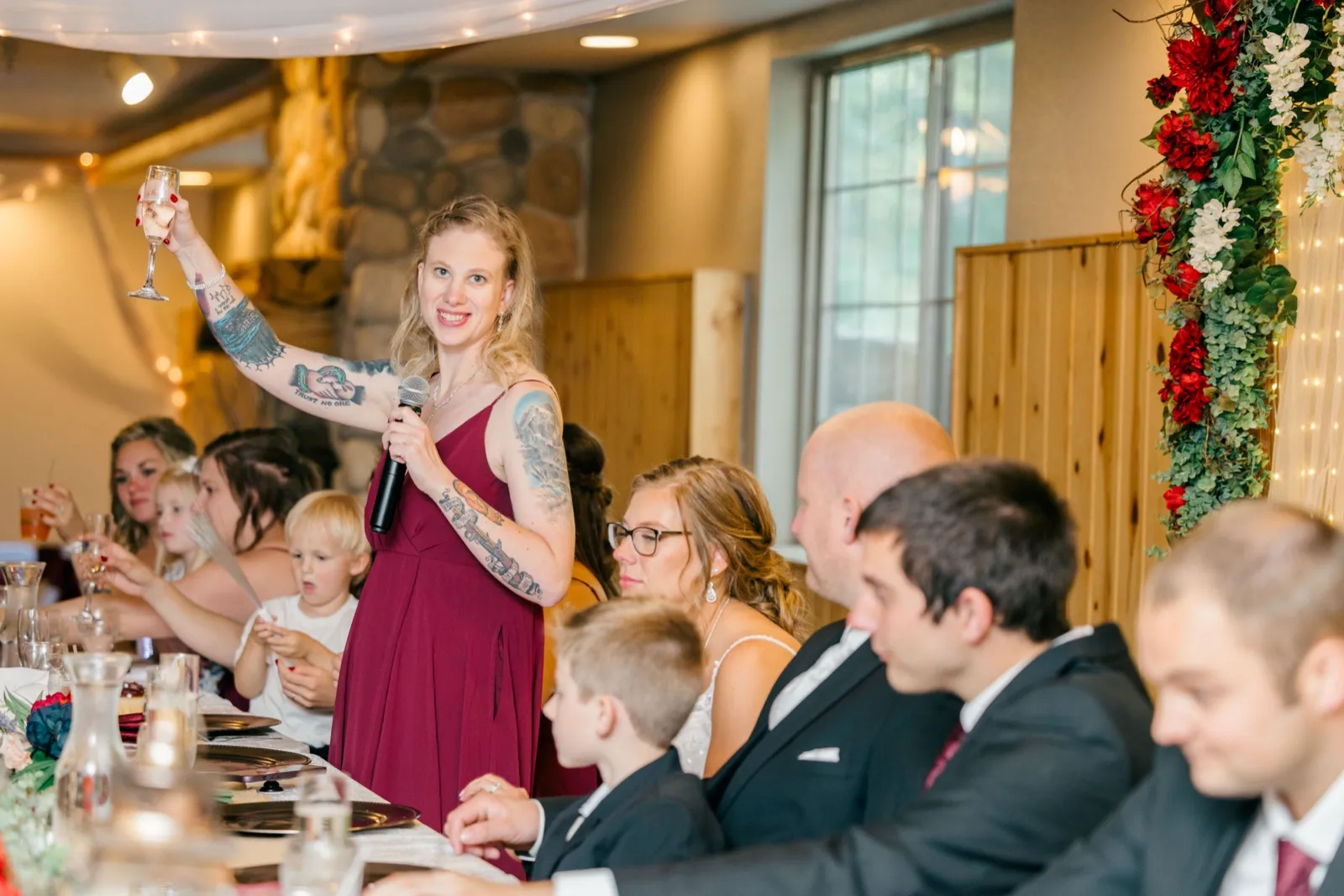 A bridesmaid in a burgundy dress raises a champagne glass while holding a microphone during a toast at the reception head table — Tim Larsen Photography, Brainerd Lakes MN